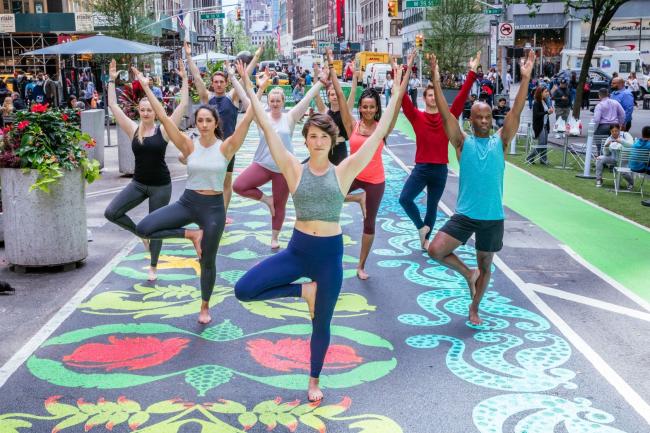 A group of 9 people standing on one leg in a yoga pose with arms up in the air while on a colorful mural in the middle of a street.