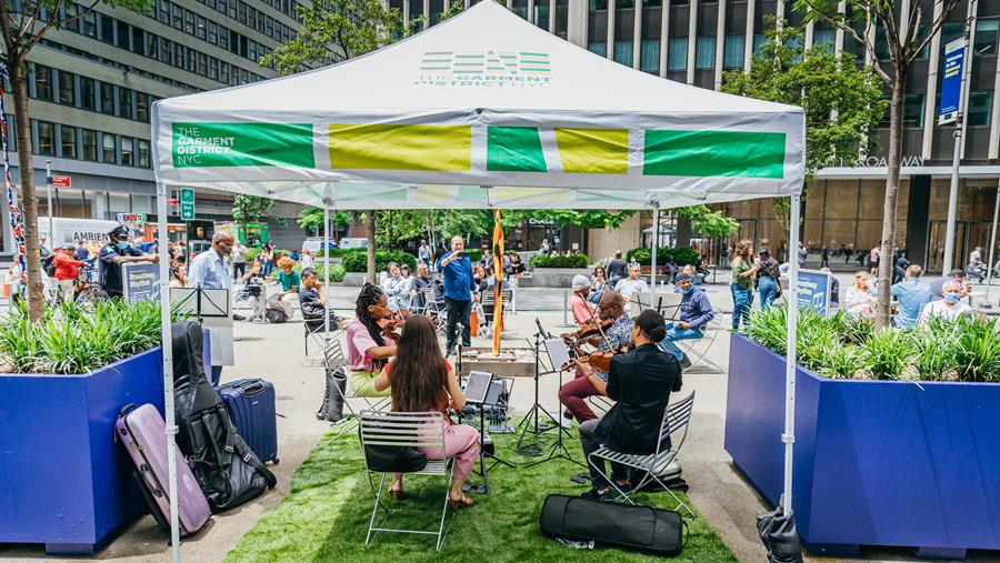 A four piece string instrument band plays under a tent in front of a crowd of people on a public plaza