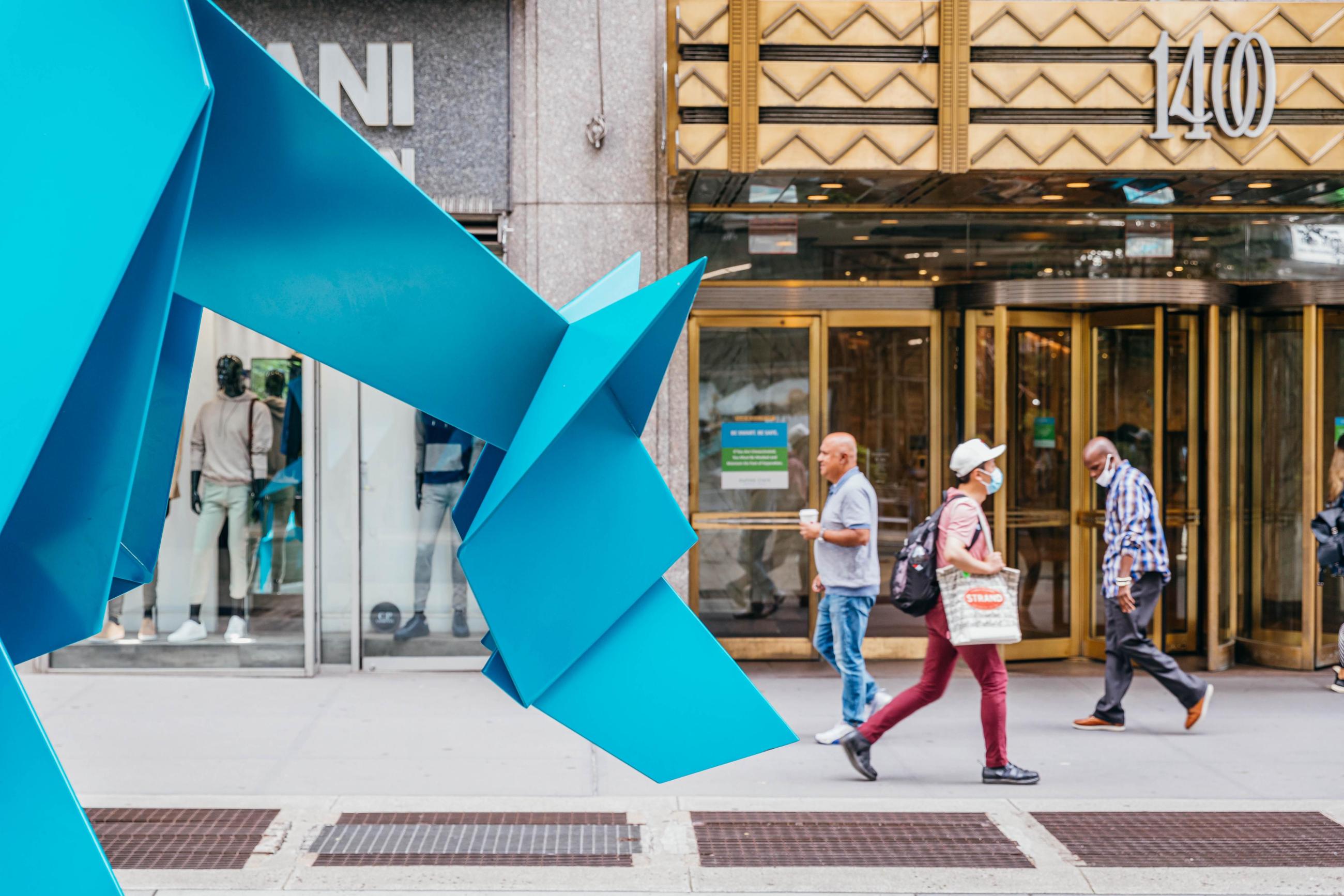 The front of a large origami blue coyote, with pedestrians walking in the background in front of a building.