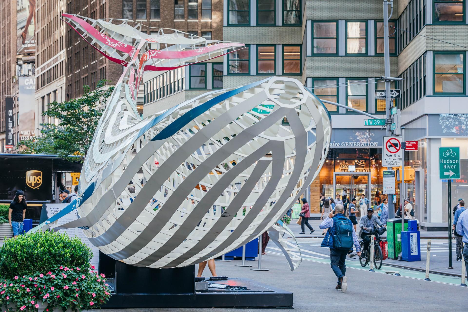 The white and blue steel mesh whale from the front angle. Pedestrians and buildings behind. 