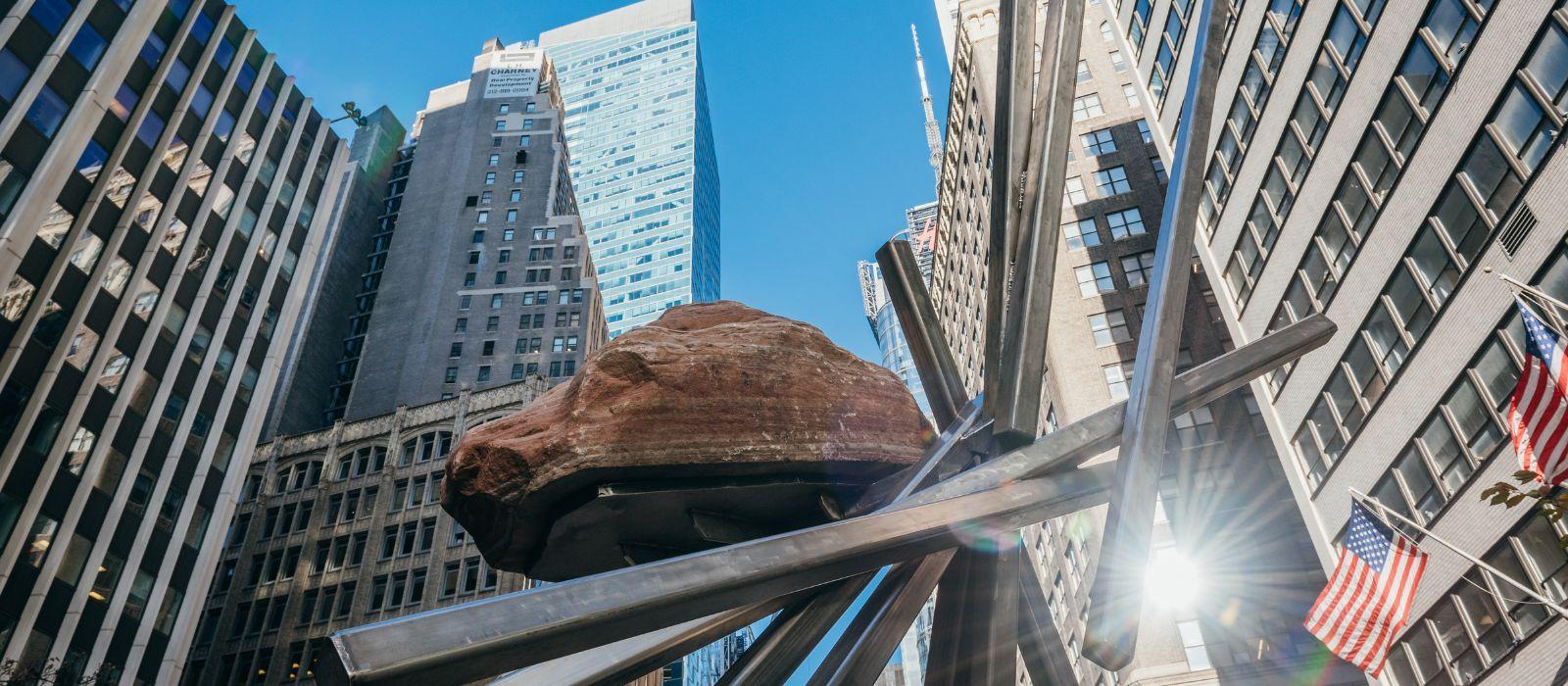 A large boulder supported by diagonal metal poles against the backdrop of skyscrapers. 