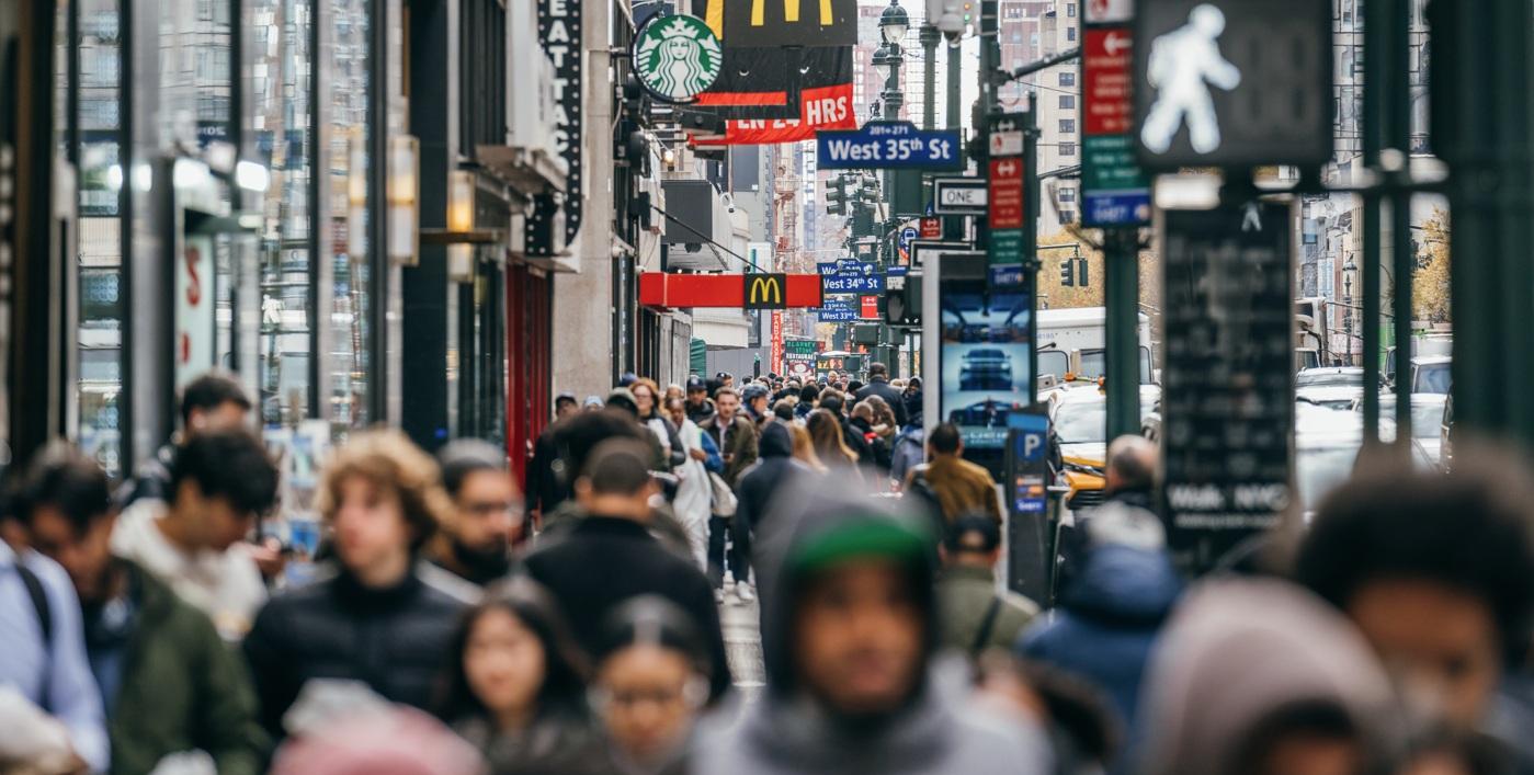 A crowd of people walking up the sidewalk of 8th avenue with signs of retail stores above them.