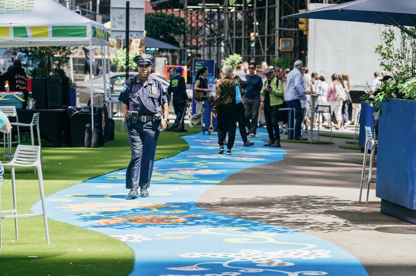 a blue parth decorated with colorful flowers curves down the middle of a pedestrian plaza with a security guard walking towards the camera. 