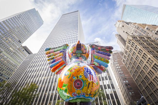 Shot from below of brightly-colored bird sculpture with skyscrapers behind it