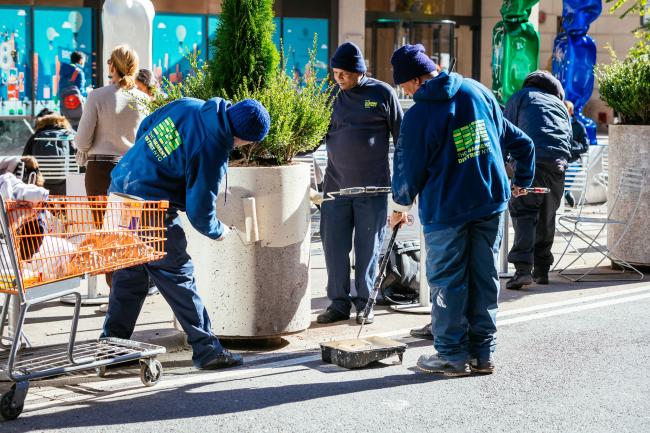 Three men in blue uniforms, painting a cement planter white.