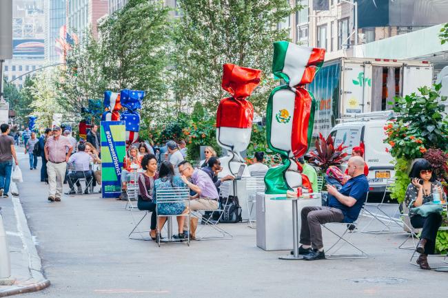 Street scene of people at tables surrounding a series of over-sized sculptures of wrapped candies