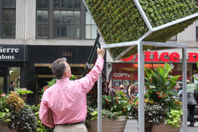 Man in a pink shirt taking a photo of cube sculpture using his cell phone