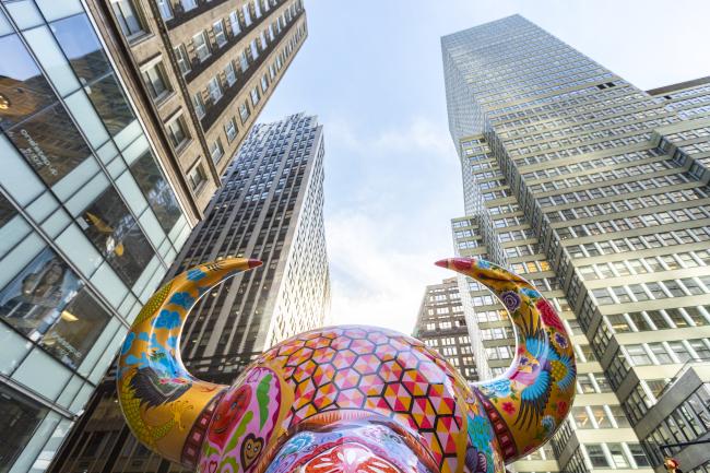 Top of the head of a brightly colored bull sculpture with skyscrapers behind