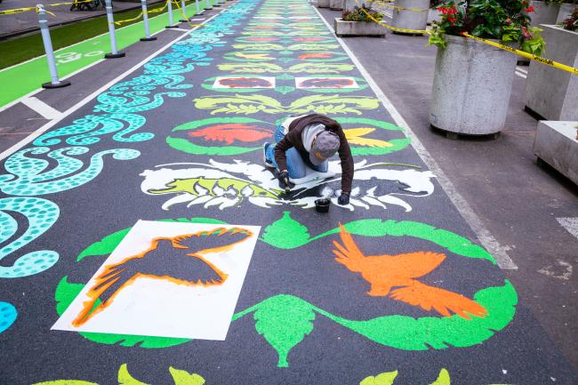 Street-level view of the artist kneeling on the mural, painting a section.