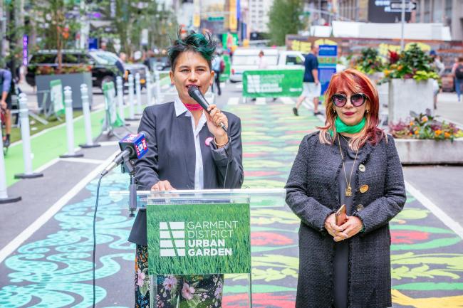 Two women at a podium in front of the street mural - one speaking into microphone.