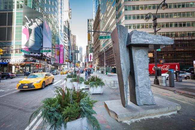 Large stone sculpture on plaza with yellow cab passing in the street alongside.