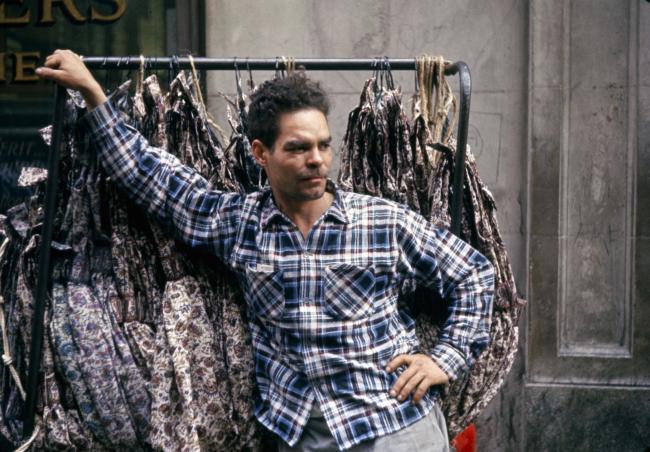 Man in a blue plaid shirt leaning against a garment rack full of clothes.