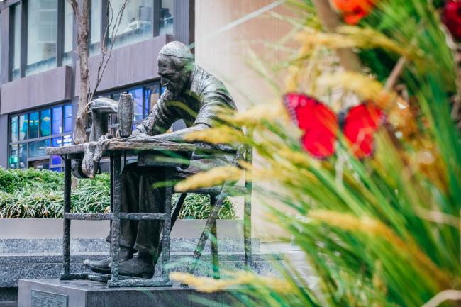 Statue of a man working at a sewing machine with decorative grasses and a red butterfly in the foreground