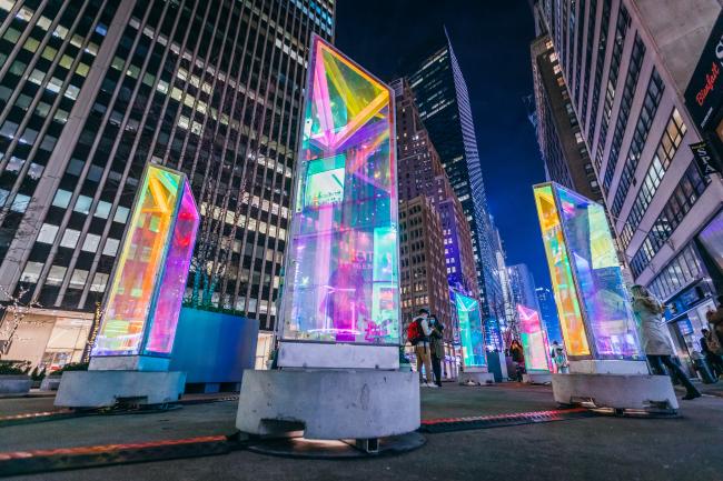 Nighttime view of five glass prism sculptures on small pedestals placed on the plaza