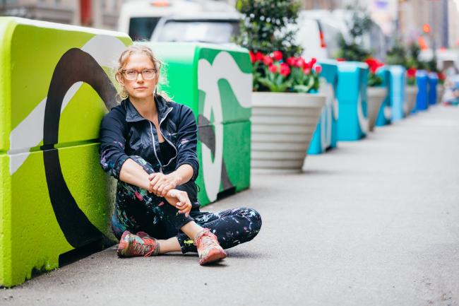 Artist Kim Carlino, in denim jacket and jeans, sits on the ground leaving against one of a column of colorful blocks running down seventh avenue.