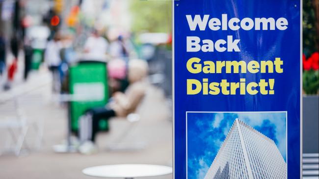 A blue sign on a public plaza that says Welcome Back Garment District