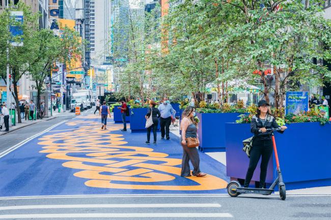 A small group of people stand on the orange and blue mural next to trees in blue plant boxes.