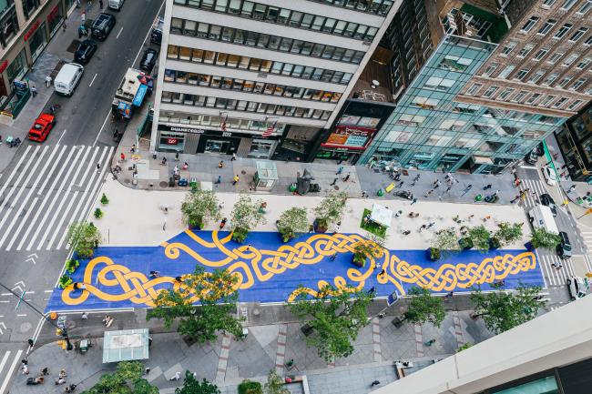 A large mural seen from above of a twist braid of orange against a deep blue cover the road on broadway.