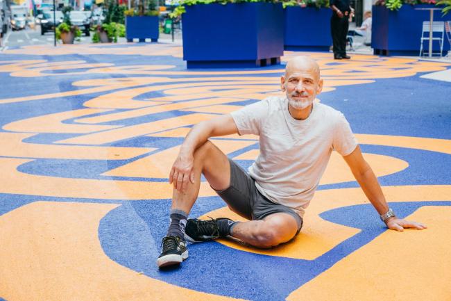Artist Steed Taylor sitting, leaning to the side on one hand, on top of his orange and blue street mural.