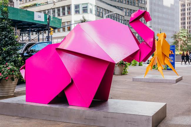 A pink elephant origami statue in the foreground on a pedestrian plaza with an orange origami animal behind it.