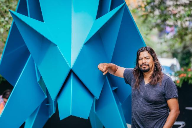 Artist Hacer with long dark hair, a short beard, and a grey tshirt, leans against his blue origami coyote statue.
