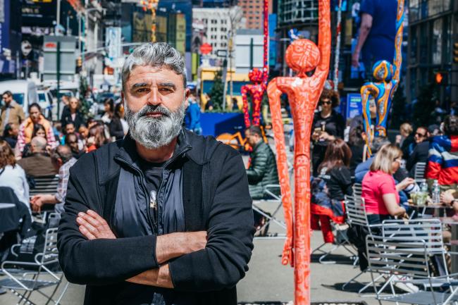 Artist Santi Flores standing with arms folded in front of a plaza full of people and his sculptures. 