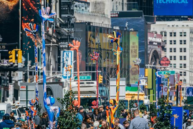 A crowded pedestrian plaza featuring colorful thin human looking statues with very tall raised hands. 