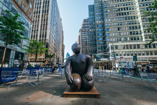 A large bronze, mostly featureless sculpture kneels in the canyon of buildings on broadway