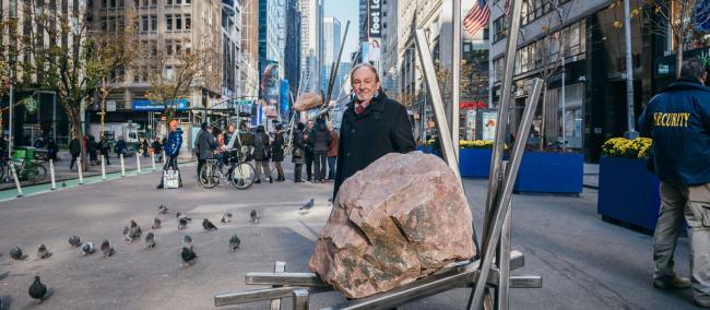 artist Del Geist standsbehind a smaller sculpture featuring a large boulder supported by diagonal metal poles