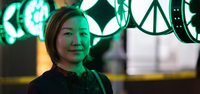 Artist Xin song smiling slightly in front of several of her green lanterns illuminating her face in light.
