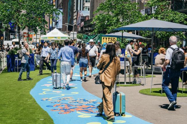 a varierty of people walk down the blue painted mural away from the camera on a sunny pedestrian plaza.