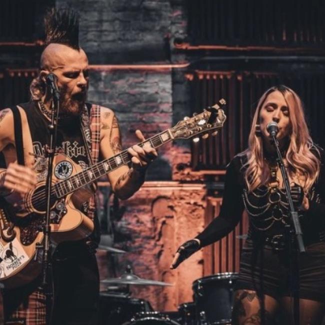 A man with a large mohawk plays the guitar while a woman in a skeletal dress sings