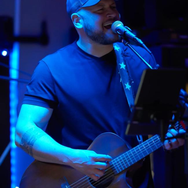 Blue Light shines on a man in a baseball cap playing guitar and singing.